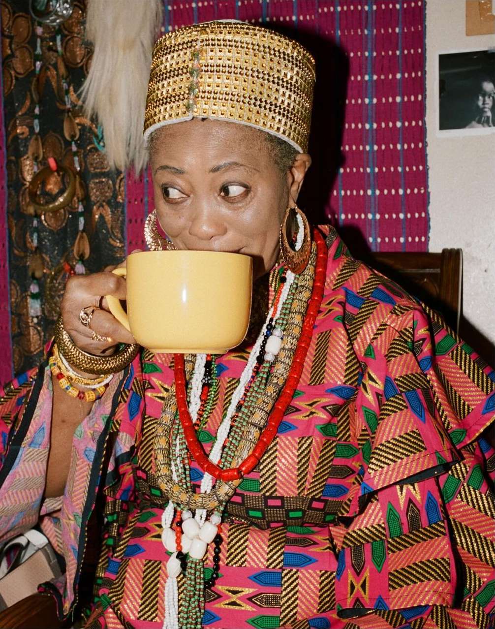Mature adult drinks from a yellow mug, wearing a patterned dress, gold headdress, and beaded jewelry inside a cozy room.