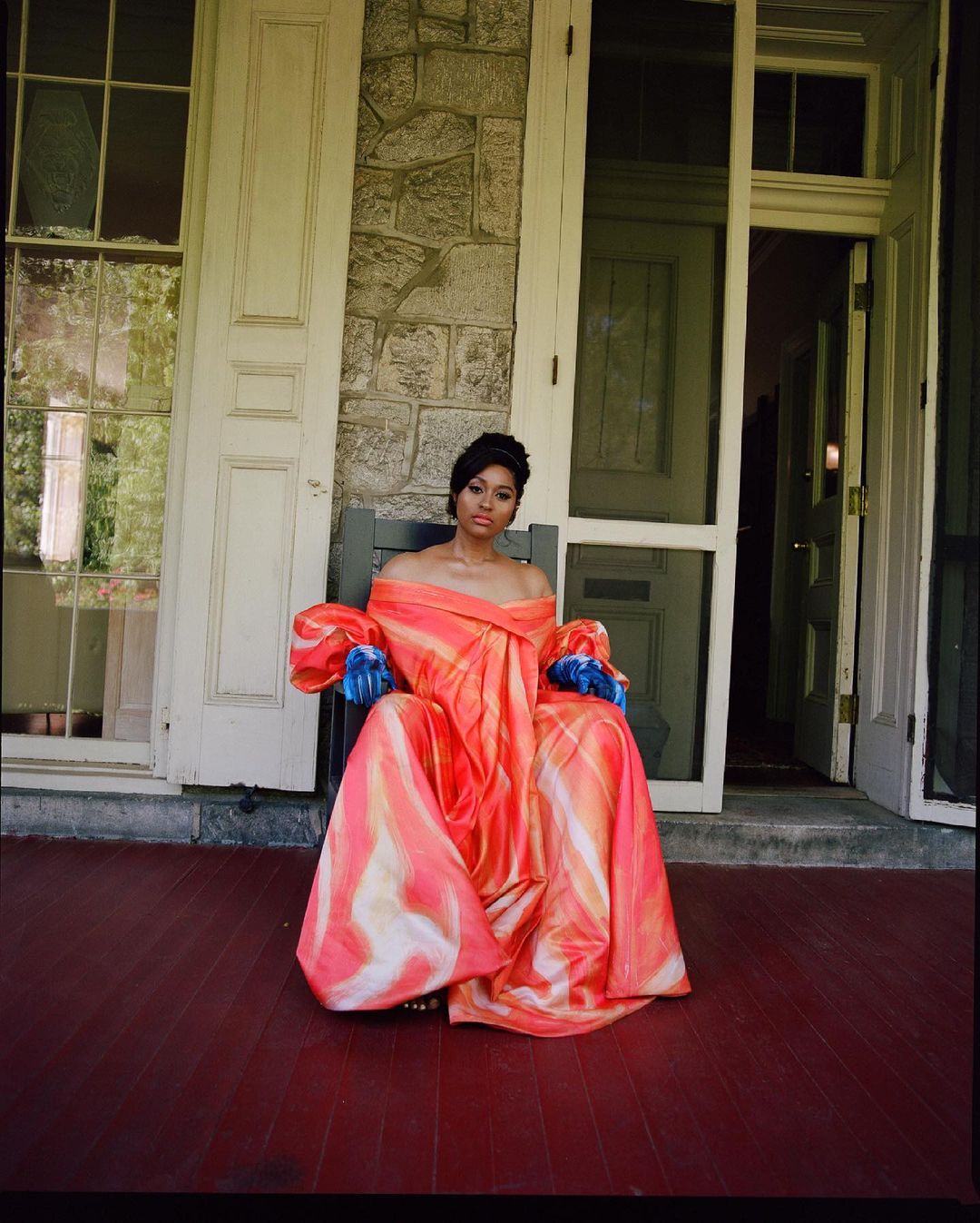 Woman in an off-the-shoulder gown sits on a porch in front of stone building with a door, surrounded by architectural details.