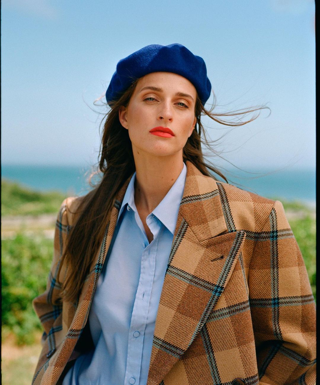 Woman in blue beret and plaid blazer posing outdoors with ocean backdrop, styled in a close-up portrait shot with soft lighting.
