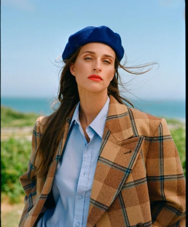 Woman in blue beret and plaid blazer posing outdoors with ocean backdrop, styled in a close-up portrait shot with soft lighting.