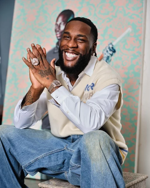 Close-up of Burna Boy smiling in an indoor setting with an art piece in the background, wearing a cream vest, jeans, and jewelry.