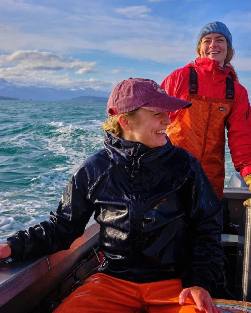 Two smiling women on a boat wearing waterproof clothing, with mountains visible in the distance under a cloudy blue sky.