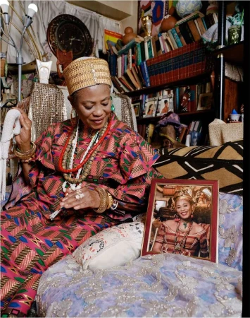 Elderly African woman sits indoors in a colorful dress and hat with jewelry, near books, a framed photo, and patterned textiles.