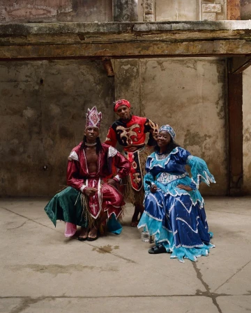 Three people in colourful costumes pose in an urban setting. One person is kneeling in the foreground, and two people are standing in the background.