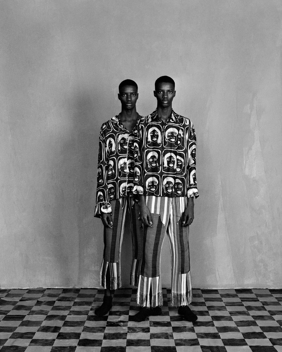 Two Black men stand side-by-side in a photo studio. They are wearing matching patterned shirts, striped pants, and black shoes.