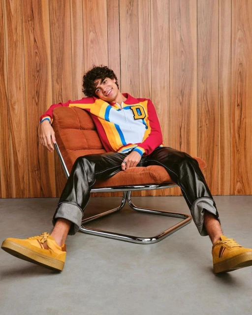 Young adult with curly hair is relaxing on a chair. He is wearing a colorful shirt and black vinyl pants with mustard color sneakers.