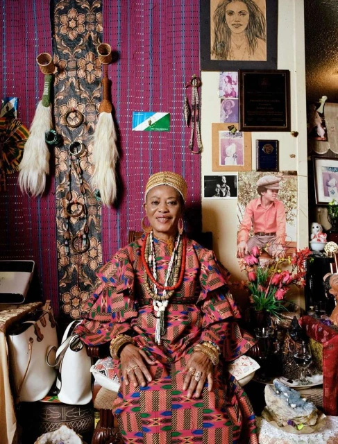 Portrait of an adult woman in a colorful patterned dress sitting in a room with a decorated wall and various objects and artworks.