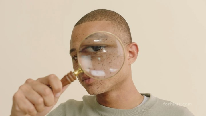 Man looks through magnifying glass held in front of his face, close-up on eye and the freckles on his cheek, neutral background.
