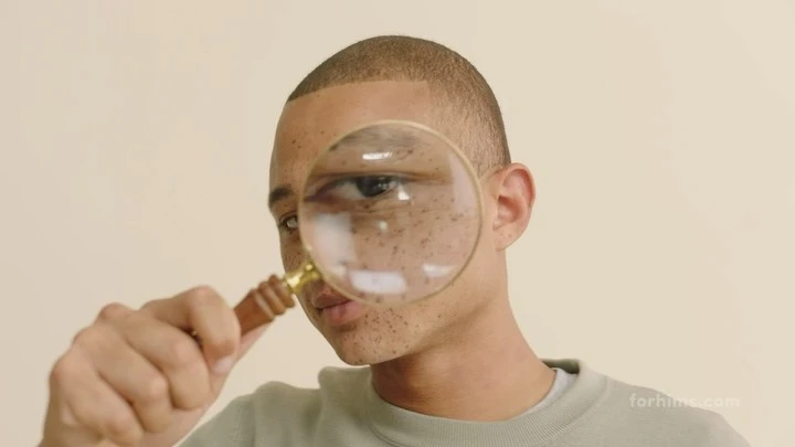 Man looks through magnifying glass held in front of his face, close-up on eye and the freckles on his cheek, neutral background.
