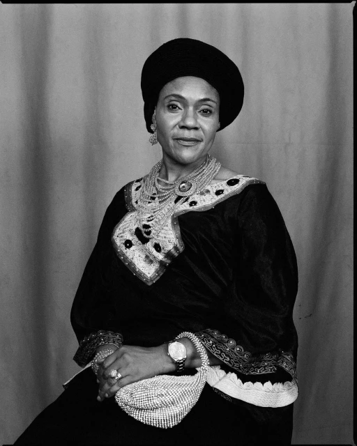Portrait of an elegant African American woman wearing a black dress, large beaded necklace, hat, and a beaded purse in a studio setting.