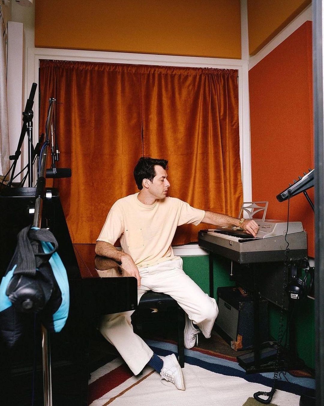 Young man in a peach shirt sits at a piano in a colorful music room, playing keys with a microphone stand and amplifier nearby.