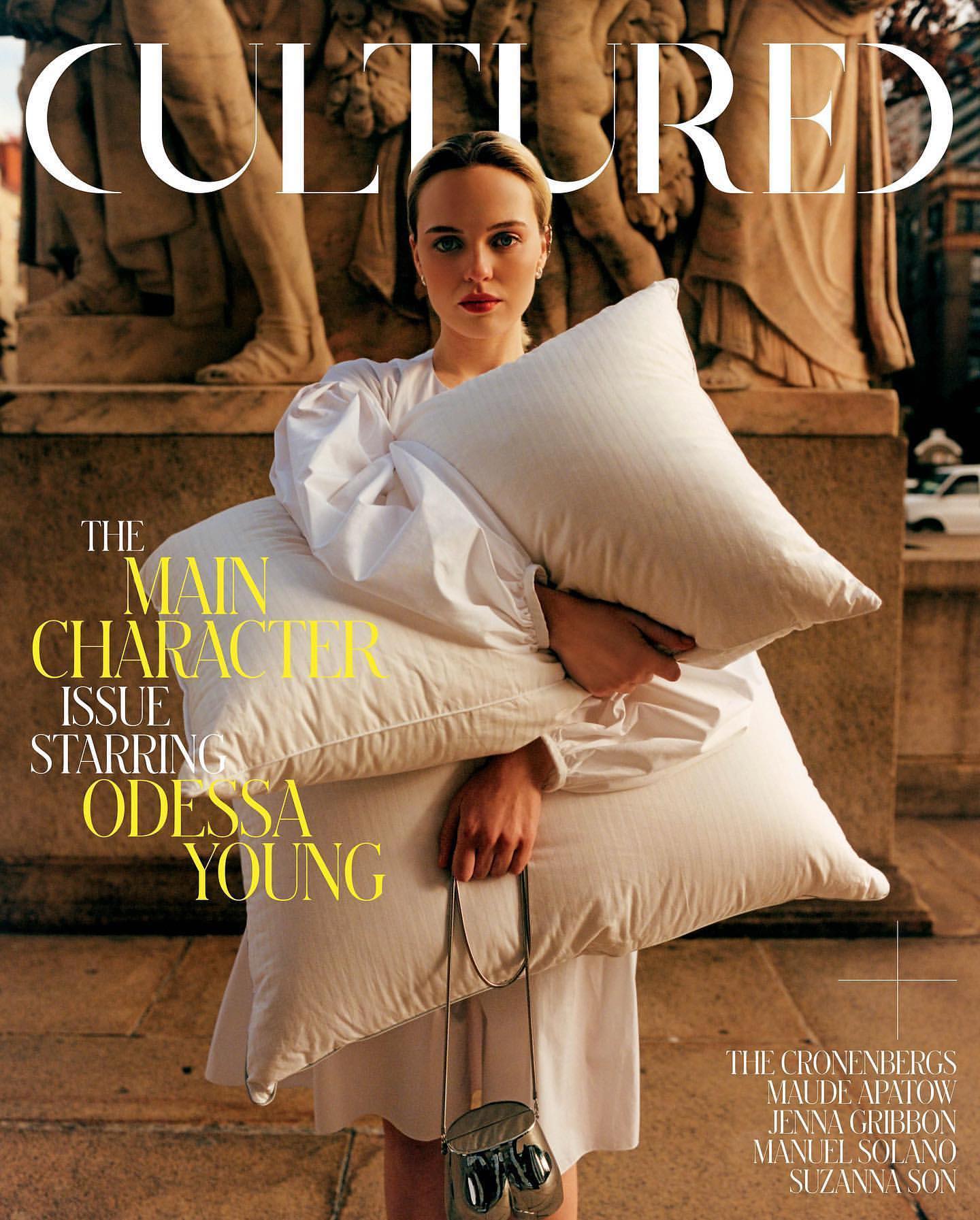 Magazine cover featuring Odessa Young holding two large pillows against a stone building facade in an outdoor setting.