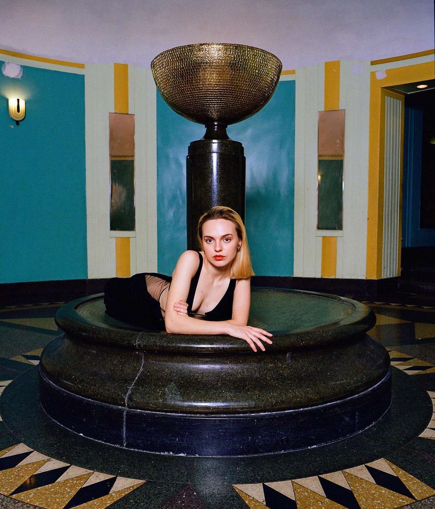 Woman in a black dress poses in a large marble fountain in an art deco style interior with gold and turquoise accents and geometric flooring.