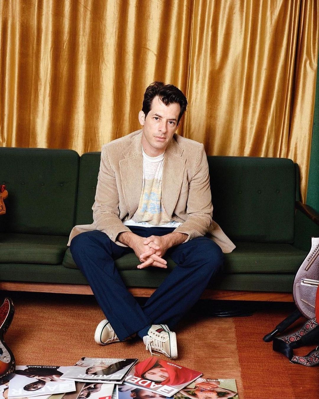 Man in a blazer seated on green couch surrounded by magazines in a brightly lit room with a gold curtain in the background.