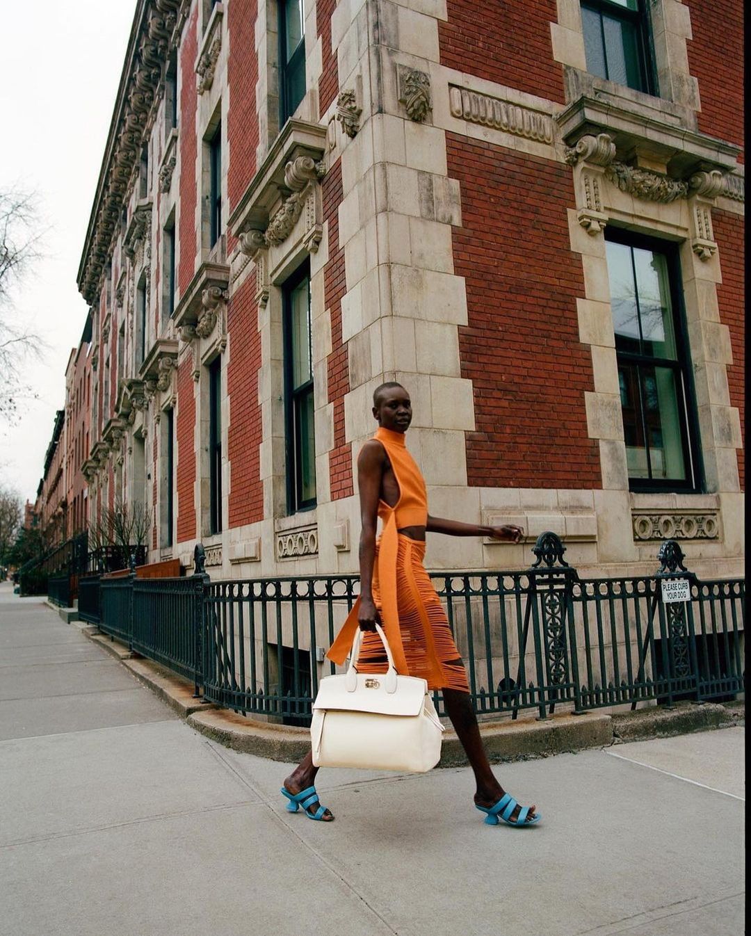 A female model walks across a city street corner in an orange dress, holding a cream-colored tote bag with brown buildings in the background.