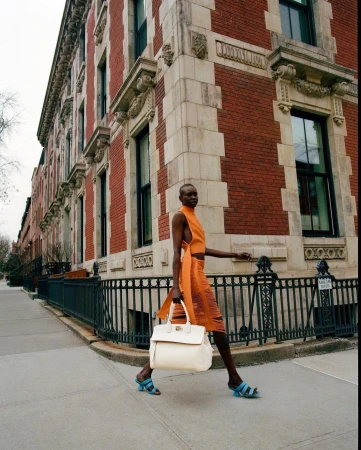 A female model walks across a city street corner in an orange dress, holding a cream-colored tote bag with brown buildings in the background.