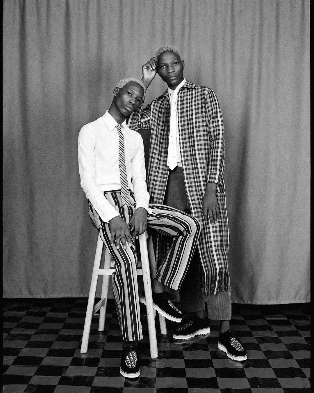 Black and white studio portrait of two blond men, one sitting on a stool and the other standing, both wearing patterned outfits.