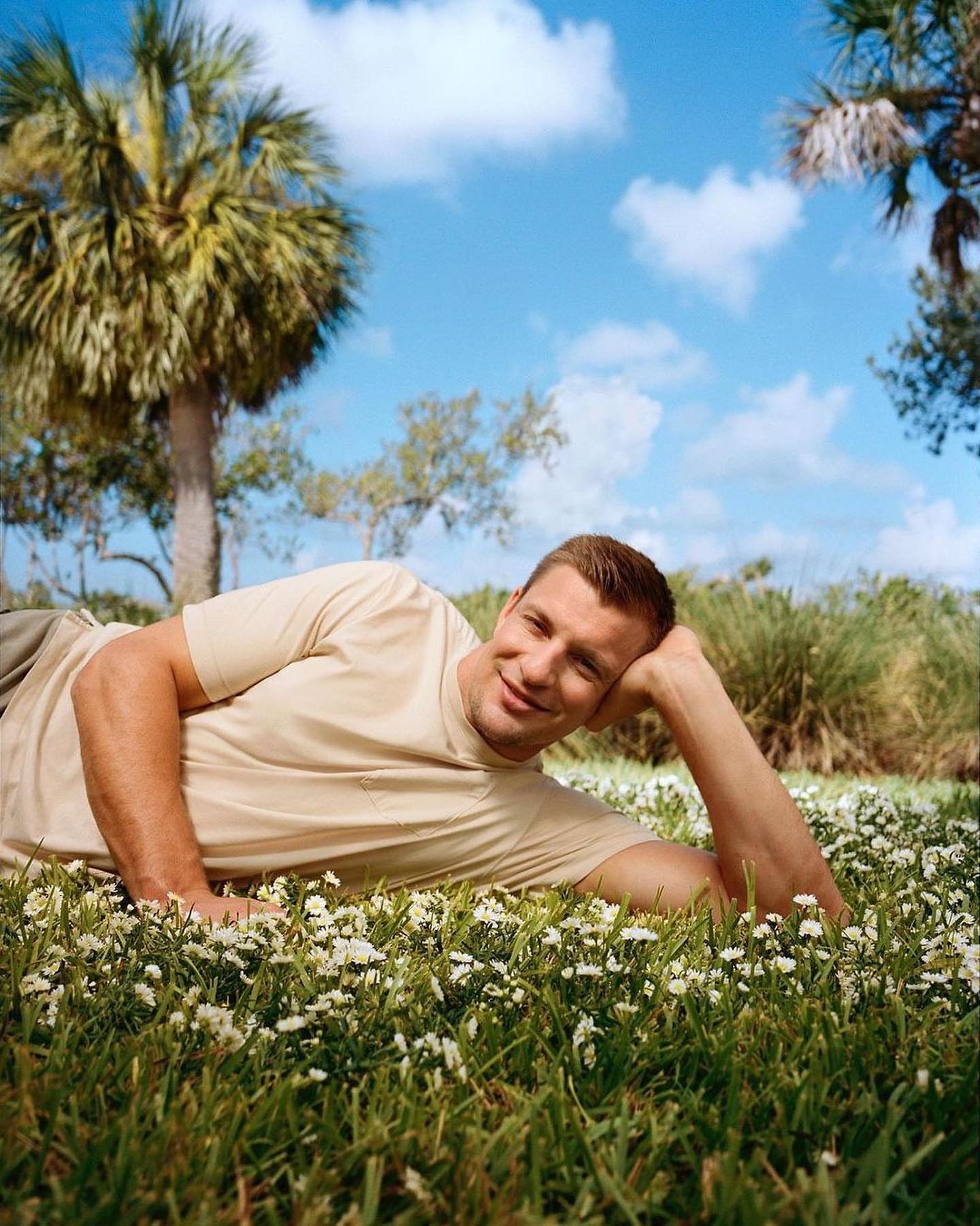 Man lying in a field of flowers, wearing a casual beige t-shirt, with green grass, palm trees and a bright blue sky in the background.