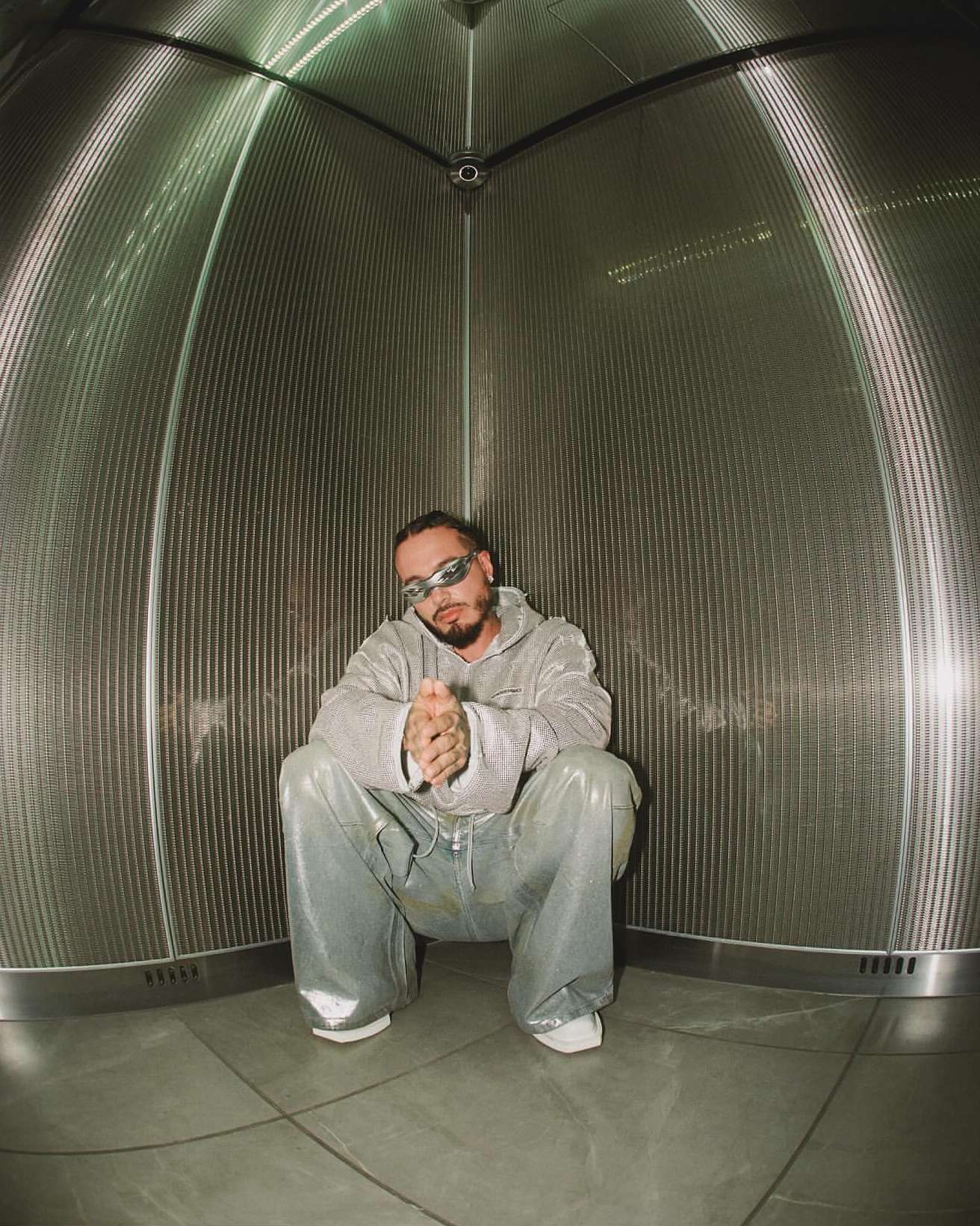 Adult male with a beard poses in a light grey hooded sweatshirt and shiny light blue pants in a metal cylindrical structure.