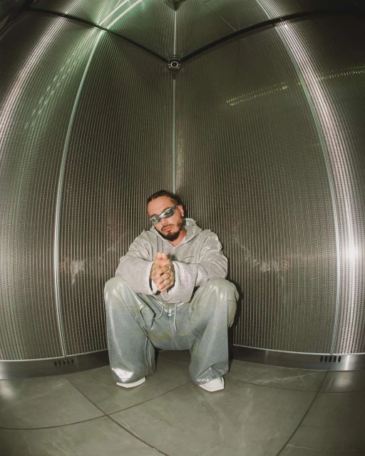 Adult male with a beard poses in a light grey hooded sweatshirt and shiny light blue pants in a metal cylindrical structure.