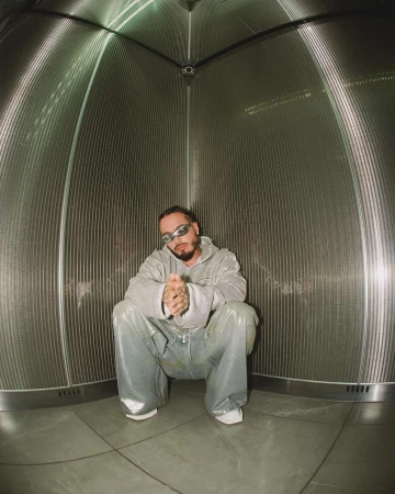 Adult male with a beard poses in a light grey hooded sweatshirt and shiny light blue pants in a metal cylindrical structure.