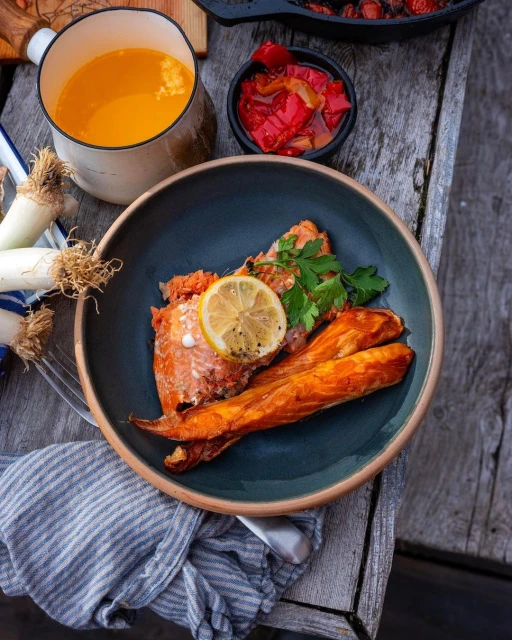 Overhead shot of cooked salmon on a plate with a lemon slice, accompanied by sweet potato, a side of peppers, and sauce.