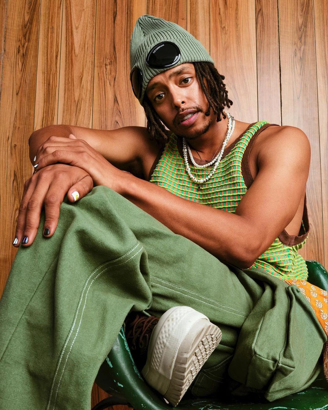 A stylish young adult male with dreadlocks, wearing a green knit beanie, tank top, and pants, sits indoors against a wood-paneled wall.