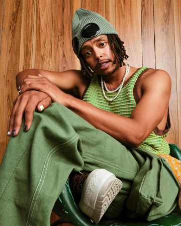 A stylish young adult male with dreadlocks, wearing a green knit beanie, tank top, and pants, sits indoors against a wood-paneled wall.