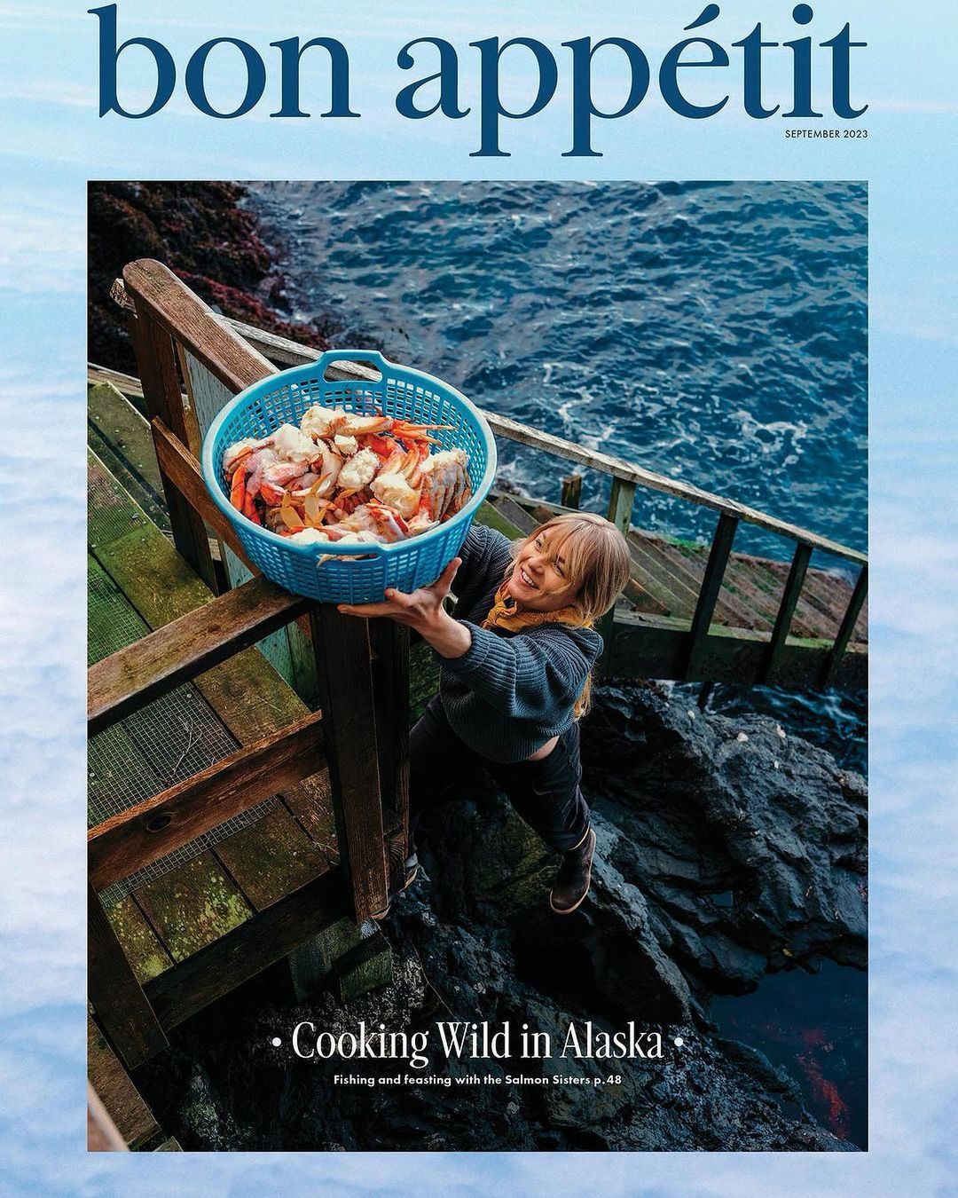 Bon Appetit magazine cover featuring a woman holding a blue basket of seafood on wooden steps near a rocky coast and ocean.