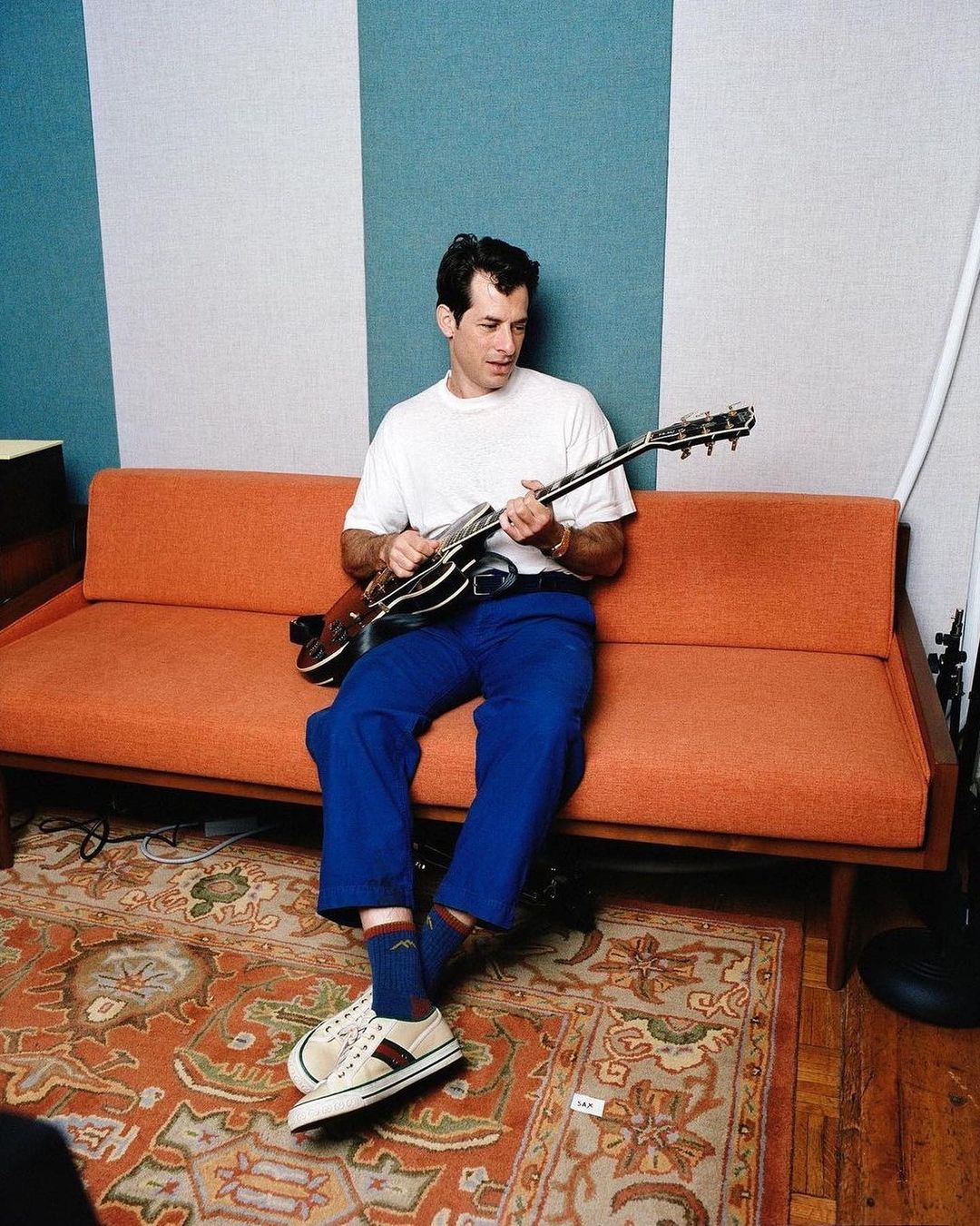 Adult male musician playing electric guitar while seated on an orange couch in a music studio with striped walls and a patterned rug.