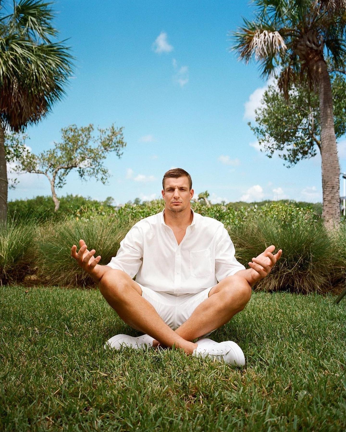 Adult male in white clothing meditates in a cross-legged pose on a green lawn with palm trees and blue sky in the background.