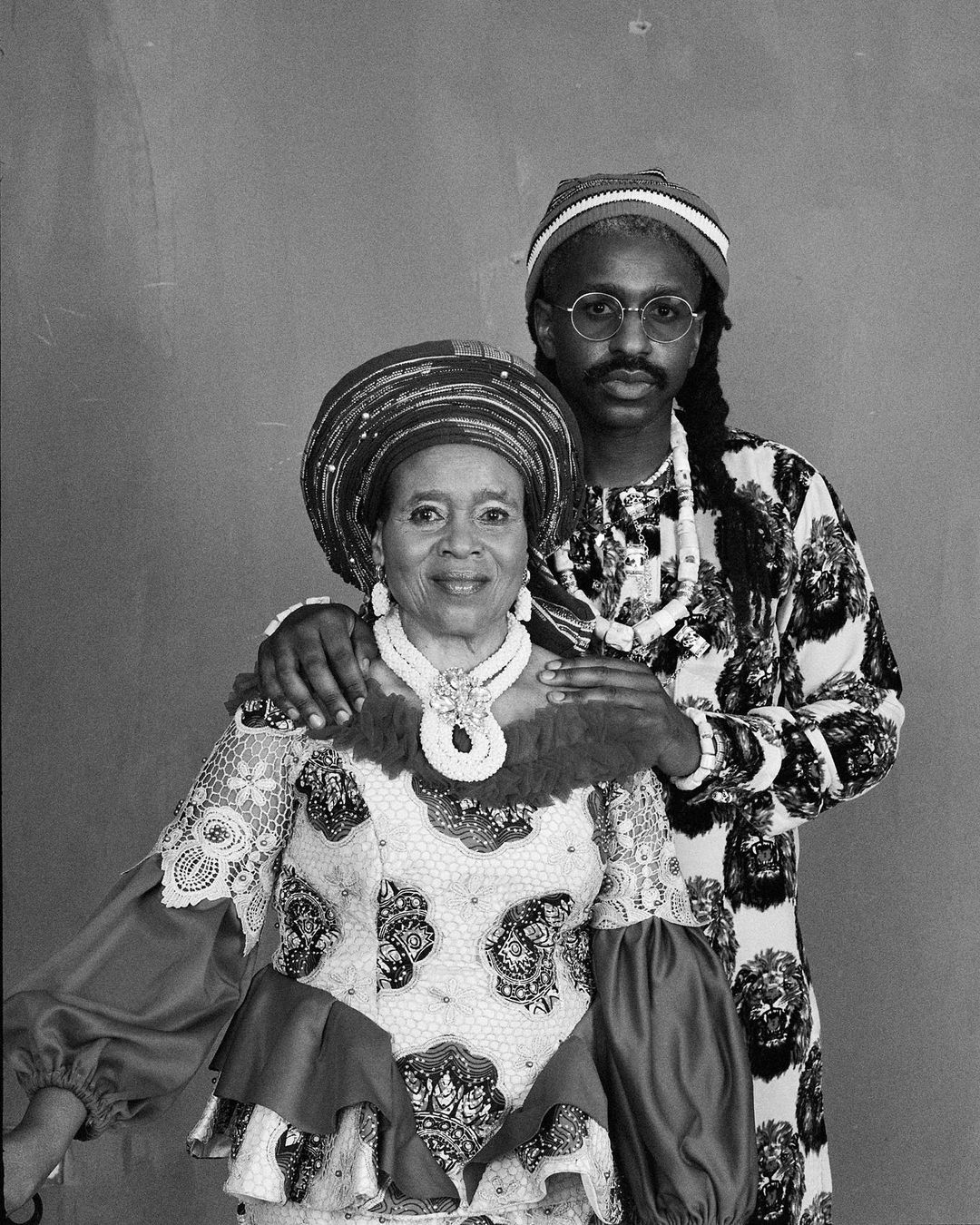 Black and white portrait of an African older adult woman and a young adult man posing together indoors, both wearing ornate traditional clothing.
