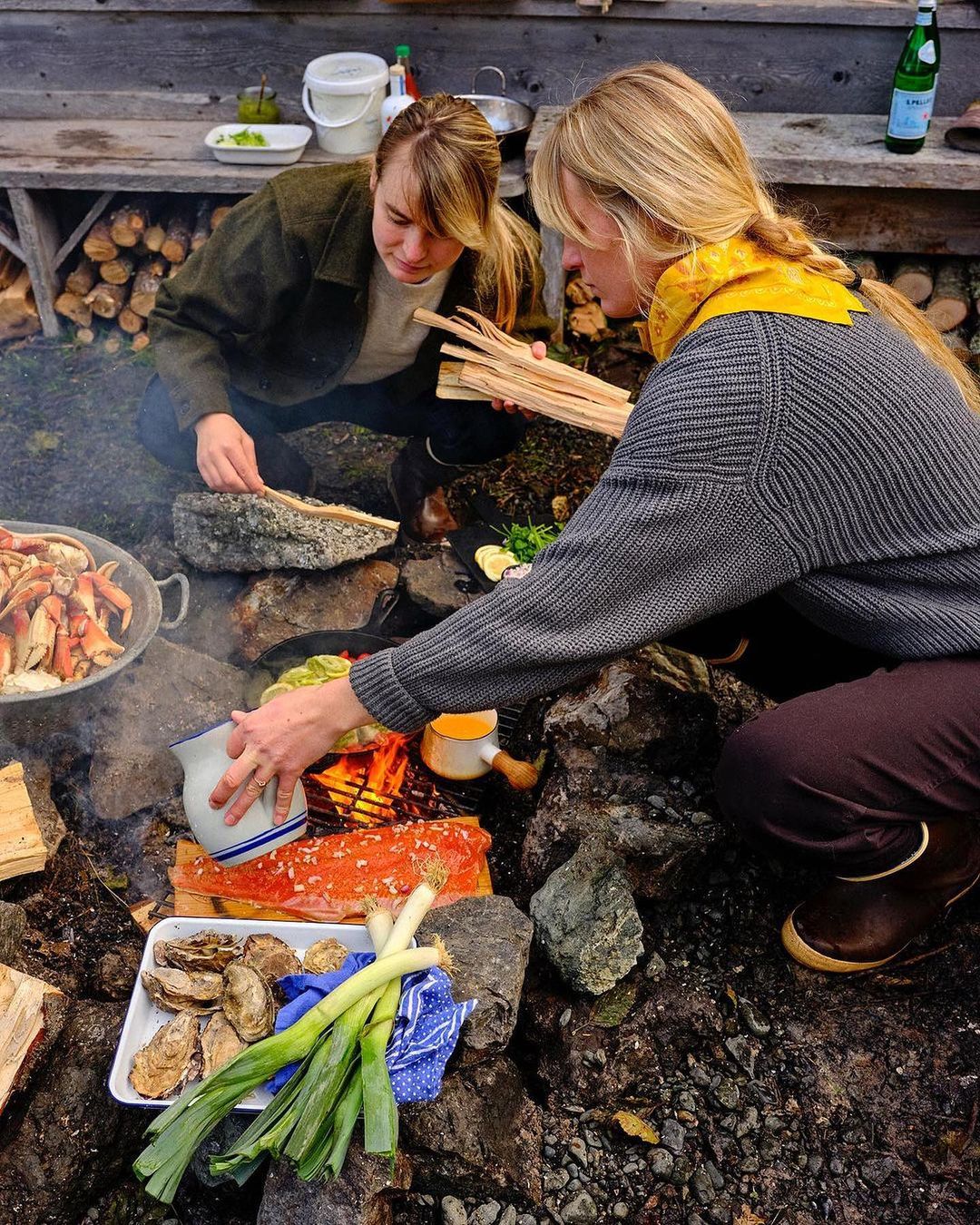 Two people prepare a meal outdoors with fresh seafood, vegetables, and fire for cooking, suggesting a culinary experience and natural setting.