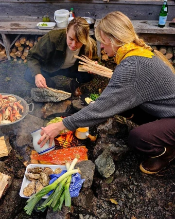 Two people prepare a meal outdoors with fresh seafood, vegetables, and fire for cooking, suggesting a culinary experience and natural setting.