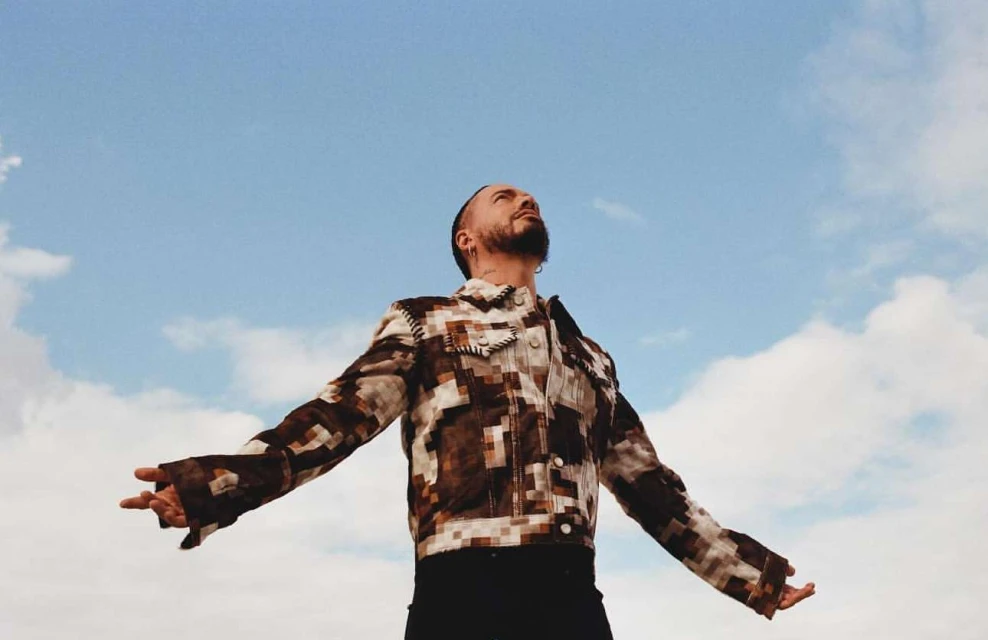 Adult male in patchwork-style jacket stands with arms open against a light blue sky with scattered clouds, photographed from a low angle.