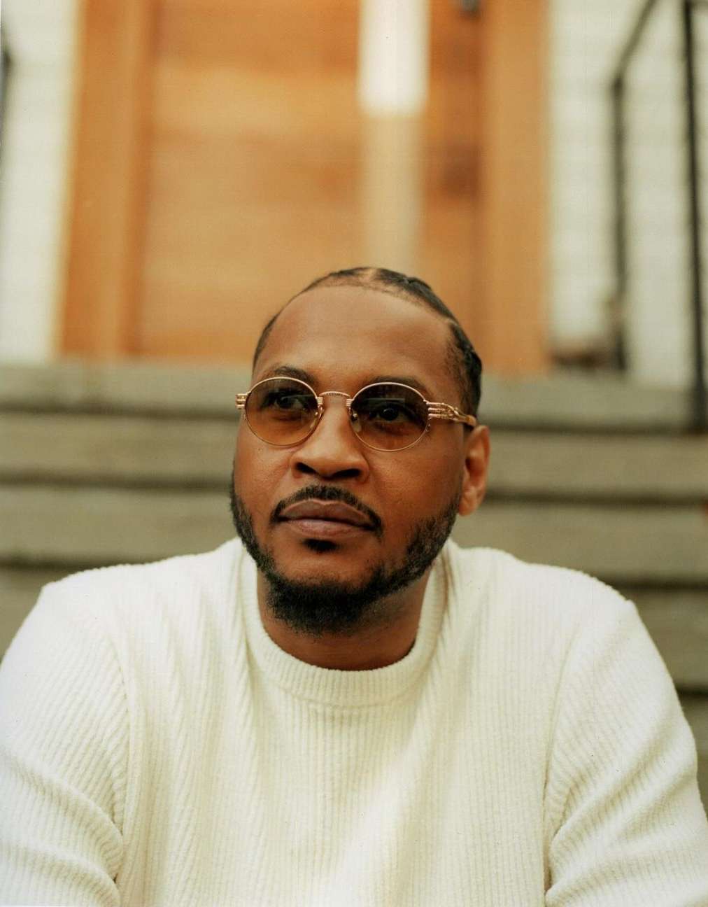 Portrait of a Black man with sunglasses wearing a white textured sweater, seated on stone steps in New York by Brad Ogbonna.