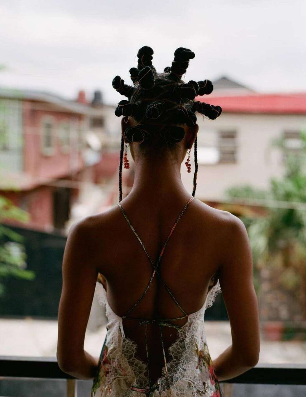 Back view of an adult woman with unique hairstyle and a floral-print dress, standing on a balcony, overlooking urban buildings.