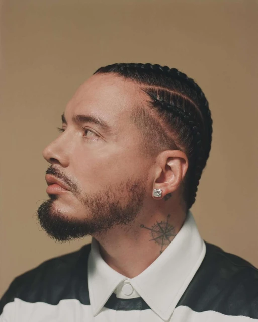 Close-up profile shot of an adult male with braided hair, a beard, and a geometric tattoo on his neck against a tan backdrop.