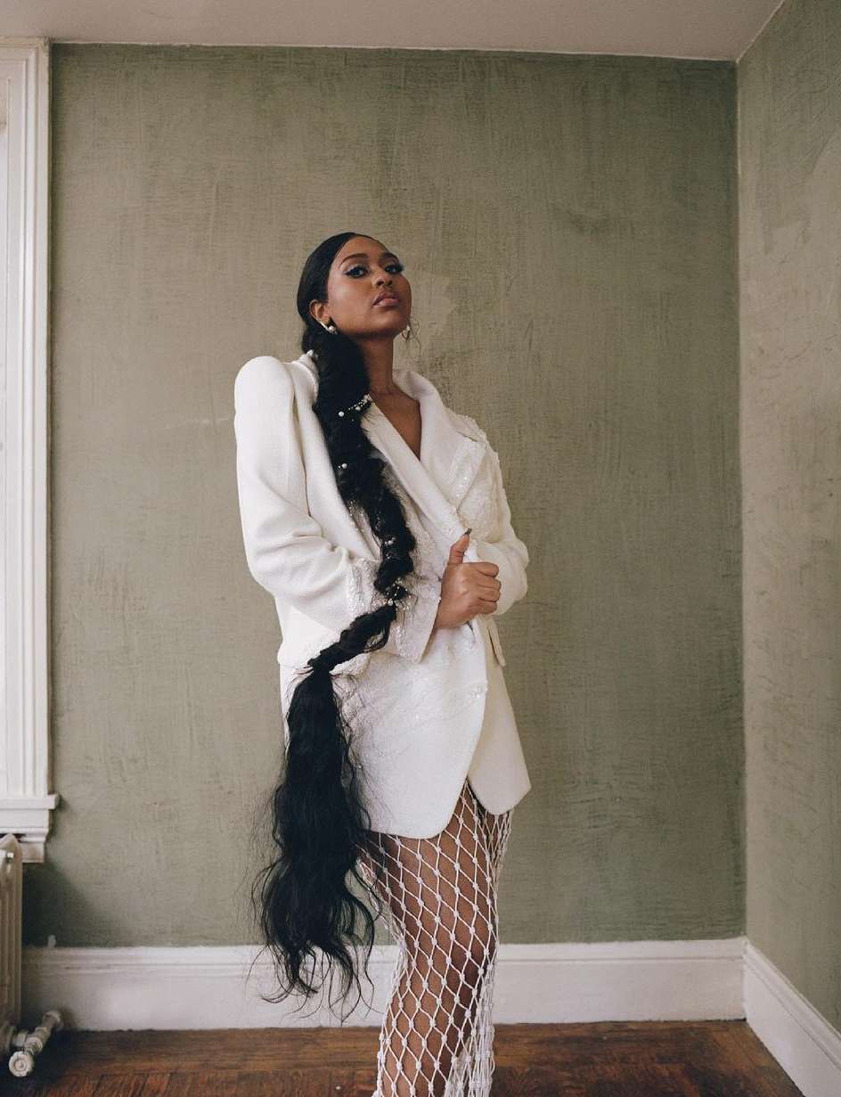 A woman with long, dark hair braided and wearing a white blazer and fishnet tights, posing in a room with textured green walls.