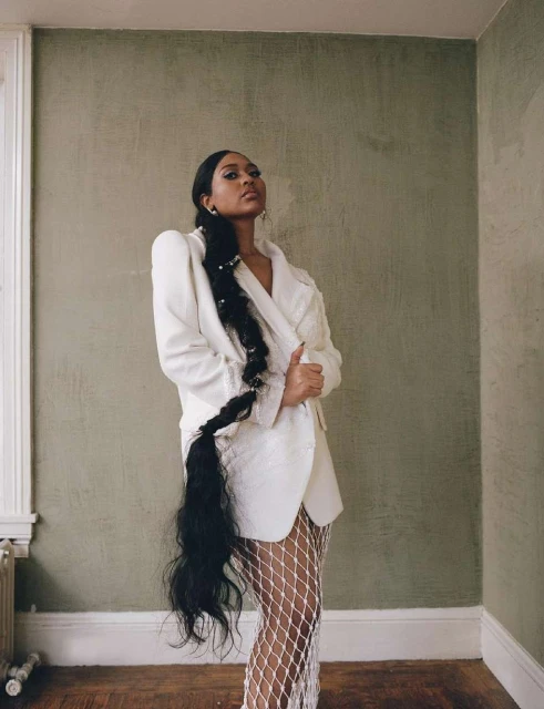 A woman with long, dark hair braided and wearing a white blazer and fishnet tights, posing in a room with textured green walls.