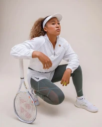 Naomi Osaka crouching in a white visor, white top, and dark grey leggings holding a tennis racket.