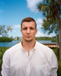 Portrait of a white man in a white button-down shirt outdoors against a body of water and palm trees under a bright blue sky.
