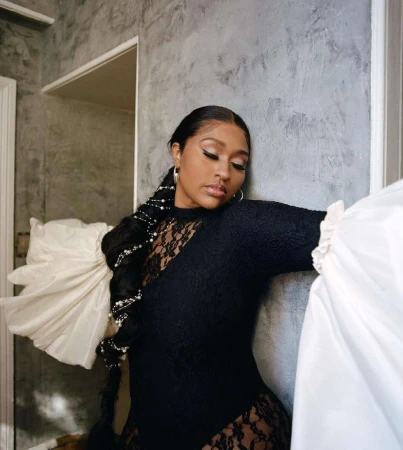 Portrait of a black woman in a black lace top with dramatic white ruffled sleeves leaning against a textured gray wall
