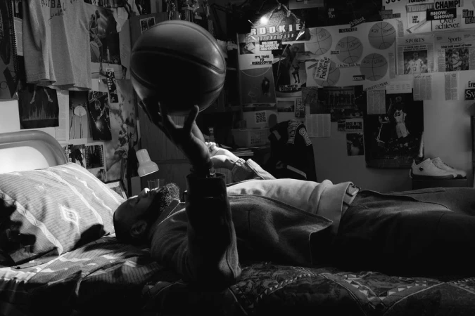 Black and white photo of a bearded adult male lying on a bed, balancing a basketball on one finger in an indoor setting.