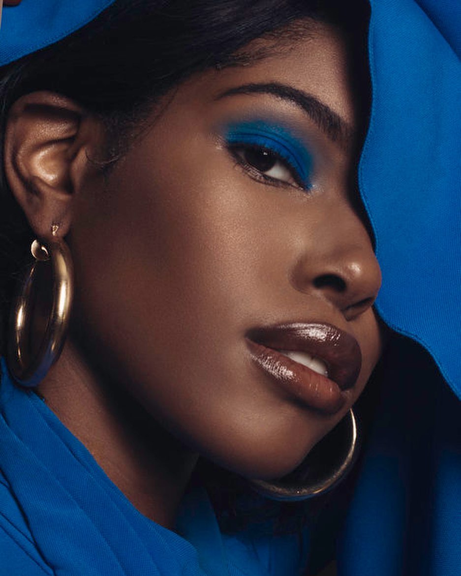 Close-up of a beautiful African-American woman with bright blue eyeshadow, glossy lips, gold hoop earrings, and a royal blue garment.