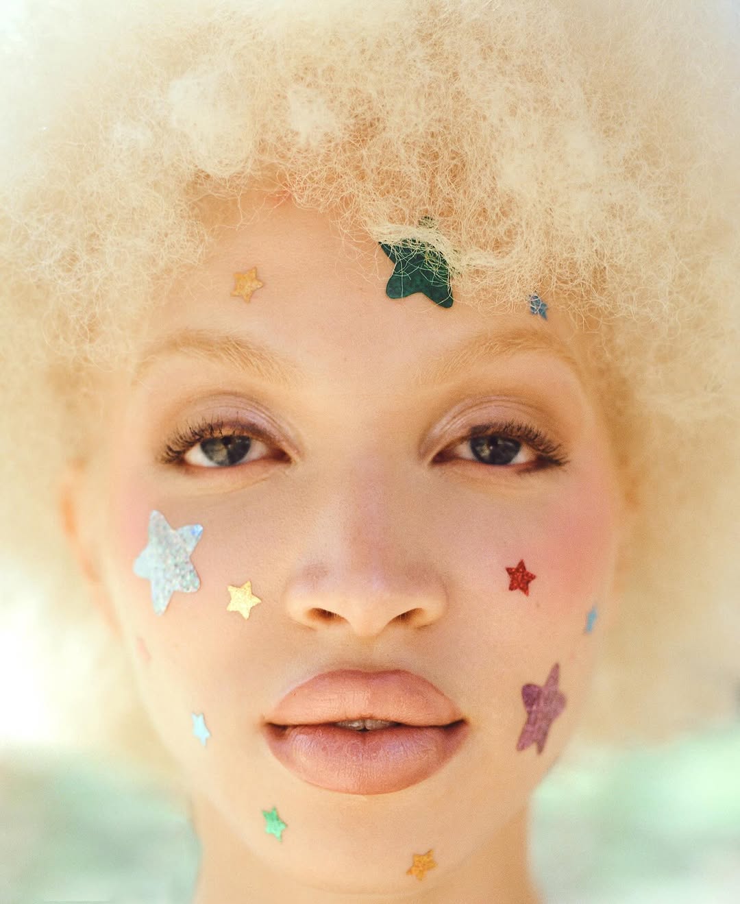 Close-up portrait of a woman with afro wearing star-shaped face gems, showcasing pastel colors and soft lighting in an outdoor setting.