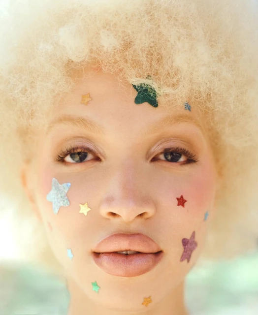 Close-up portrait of a woman with afro wearing star-shaped face gems, showcasing pastel colors and soft lighting in an outdoor setting.