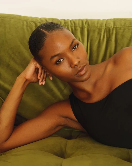 Portrait of a woman with brown skin lying on an olive green tufted sofa in a black top looking at the camera indoors.