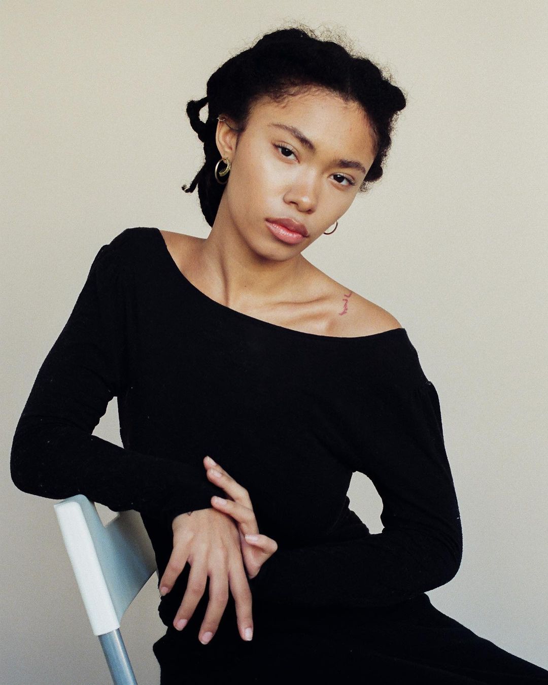 Portrait of a young black woman in a black top seated against a plain light wall, photographed by Brandon Hicks.