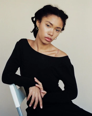 Portrait of a young black woman in a black top seated against a plain light wall, photographed by Brandon Hicks.